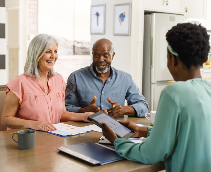 a smiling older couple sitting at a table discussing with a woman holding a tablet, cozy home setting, promoting positive communication and connection