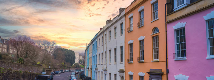 A mother carries her daughter on her shoulders, both wearing matching outfits, with a house in the background surrounded by greenery. Family bonding and home life.