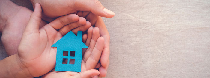A pair of hands holding a blue paper house, symbolizing home ownership and family protection.