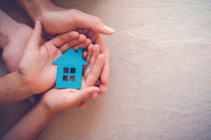 Hands of different sizes holding a blue house-shaped cutout, symbolizing family and home protection.