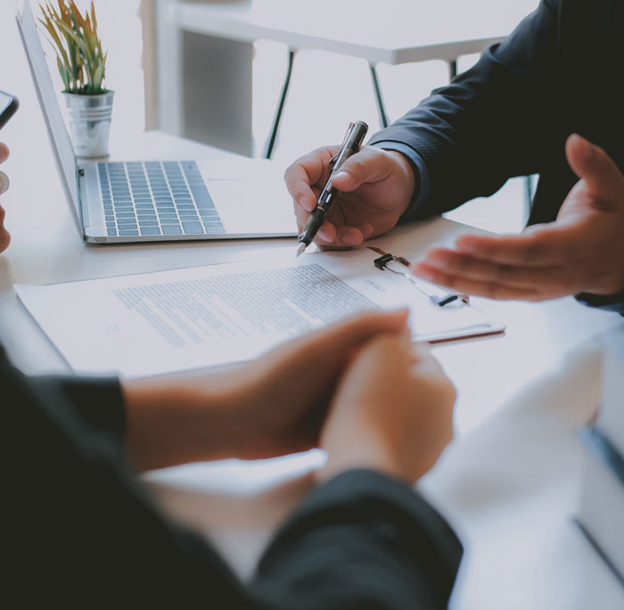 Two professionals discussing documents at a table, with a laptop and a small potted plant in the background. The scene indicates a business meeting focused on important paperwork, showcasing a professional environment.