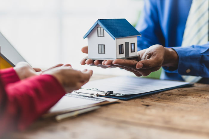 A person handing over a small model house to another individual, with documents visible on a wooden table, symbolizing a real estate transaction or house buying process.
