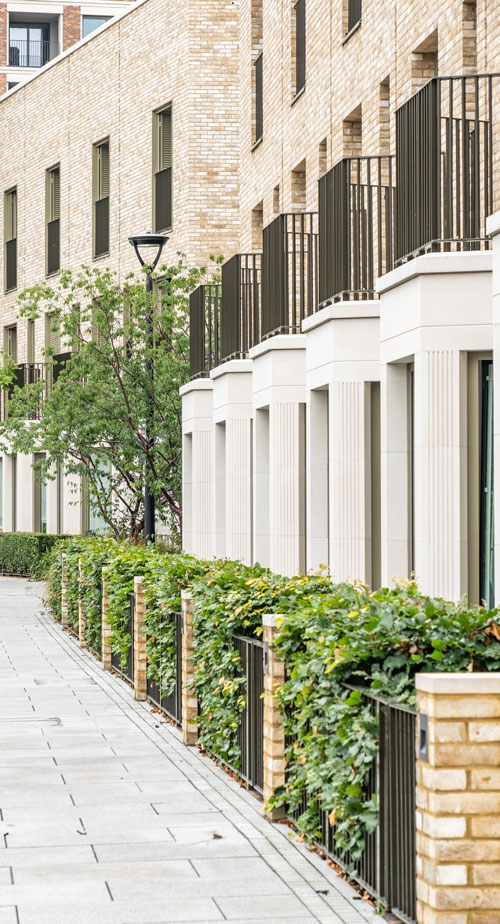 Modern residential buildings with brick facades, balconies, and lush greenery along the pathway. The image features a clean, well-maintained urban environment that emphasizes contemporary architecture and landscaping.
