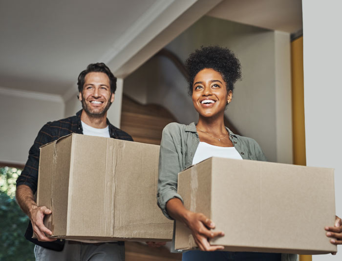 A man and a woman carrying cardboard boxes, smiling while moving into a new home.