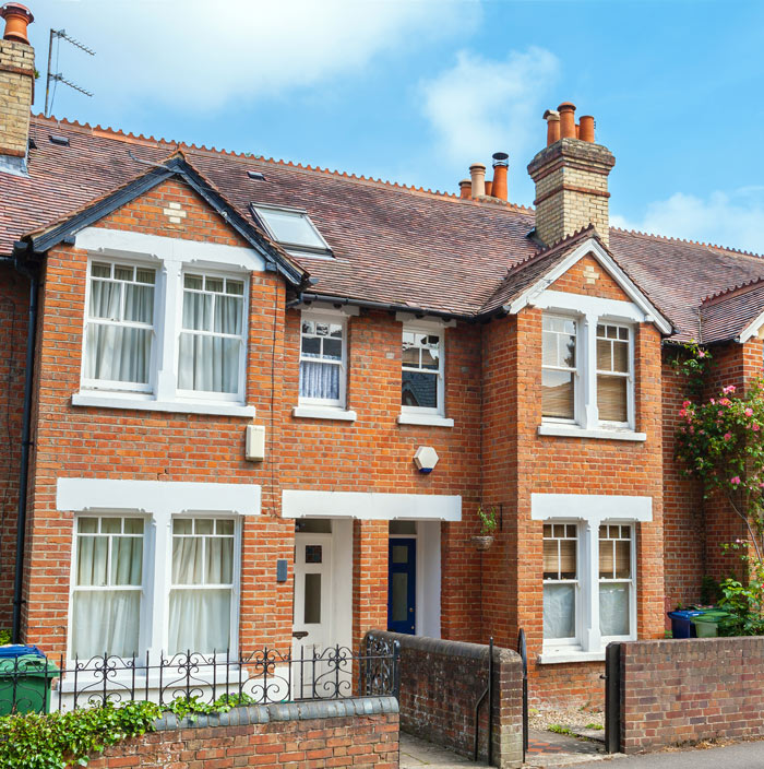 A charming brick townhouse featuring large windows, a blue door, and decorative chimney stacks, surrounded by greenery and a wrought iron fence. Ideal for cozy home living.