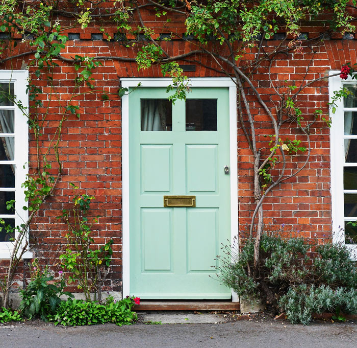 A charming mint green door set against a rustic red brick wall, surrounded by climbing plants and greenery, creating a picturesque home entrance. Perfect for home exterior inspiration.