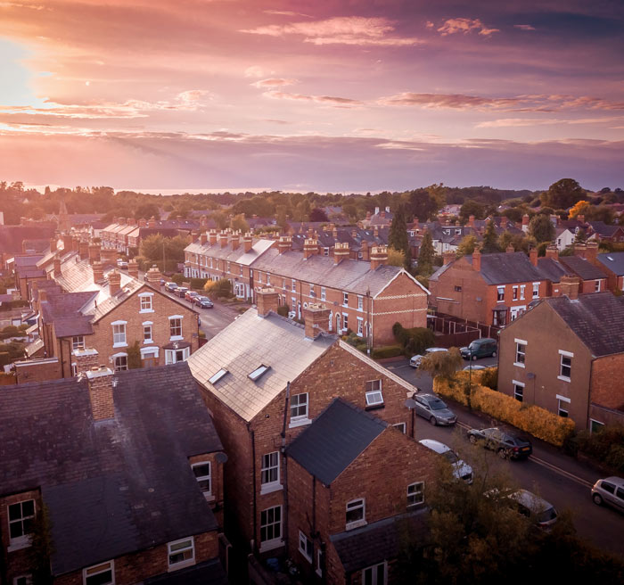Aerial view of a residential neighborhood at sunset, featuring brick houses with varying roof styles, parked cars, and lush greenery in the background.
