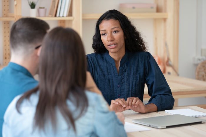 A woman with curly hair wearing a blue blouse is engaged in a conversation with two people, one facing her directly and the other partially turned, at a table with a laptop and notebook, suggesting a collaborative discussion or meeting.