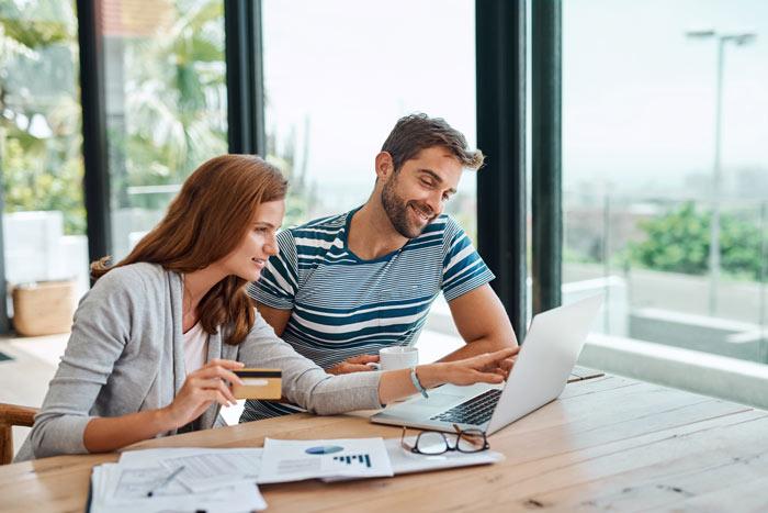 A woman holds a credit card while smiling at a man as they both look at a laptop together, with documents and glasses on the table, in a bright, modern workspace.